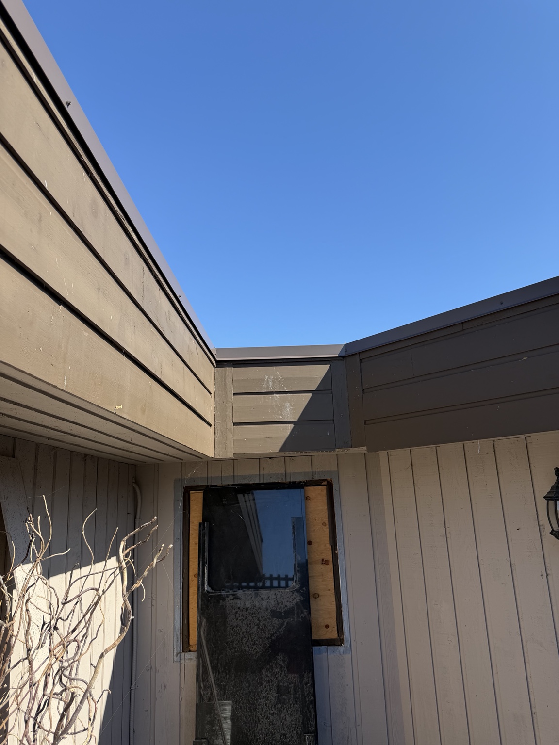 Roof edge with metal cap flashing and dark fascia above a boarded window opening