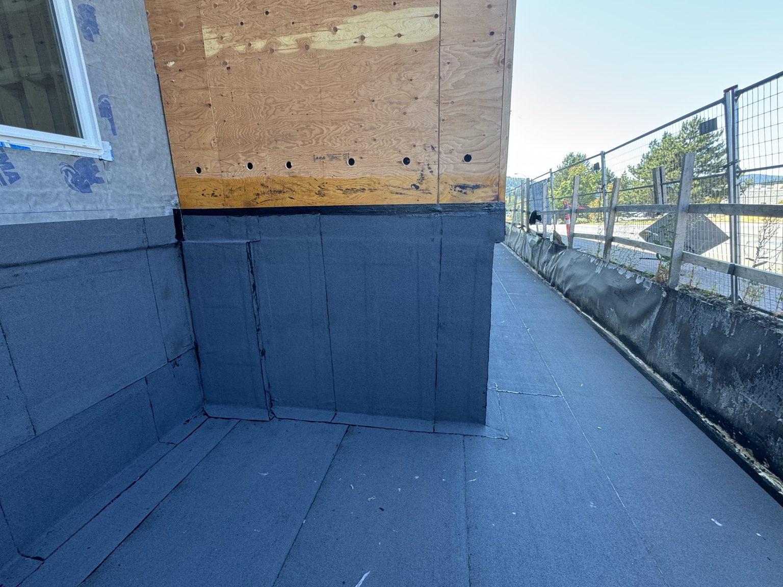 Membrane upstand with seams at a plywood-sheathed wall corner along a fenced walkway